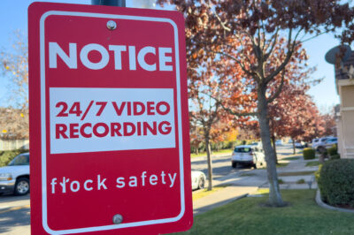 A red Flock Safety sign on a pole reads "Notice: 24/7 Video Recording" on a residential street, with parked cars, homes, trees, and a clear blue sky in the background.