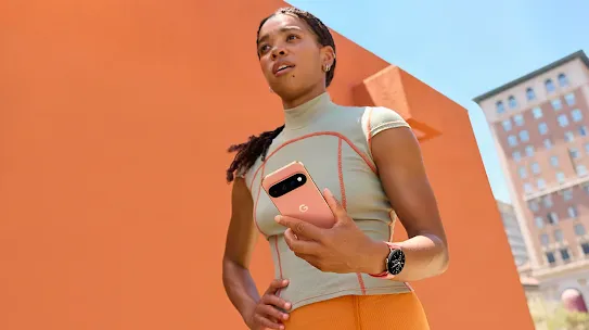 A woman outdoors holding a pink Pixel phone against an orange building background.